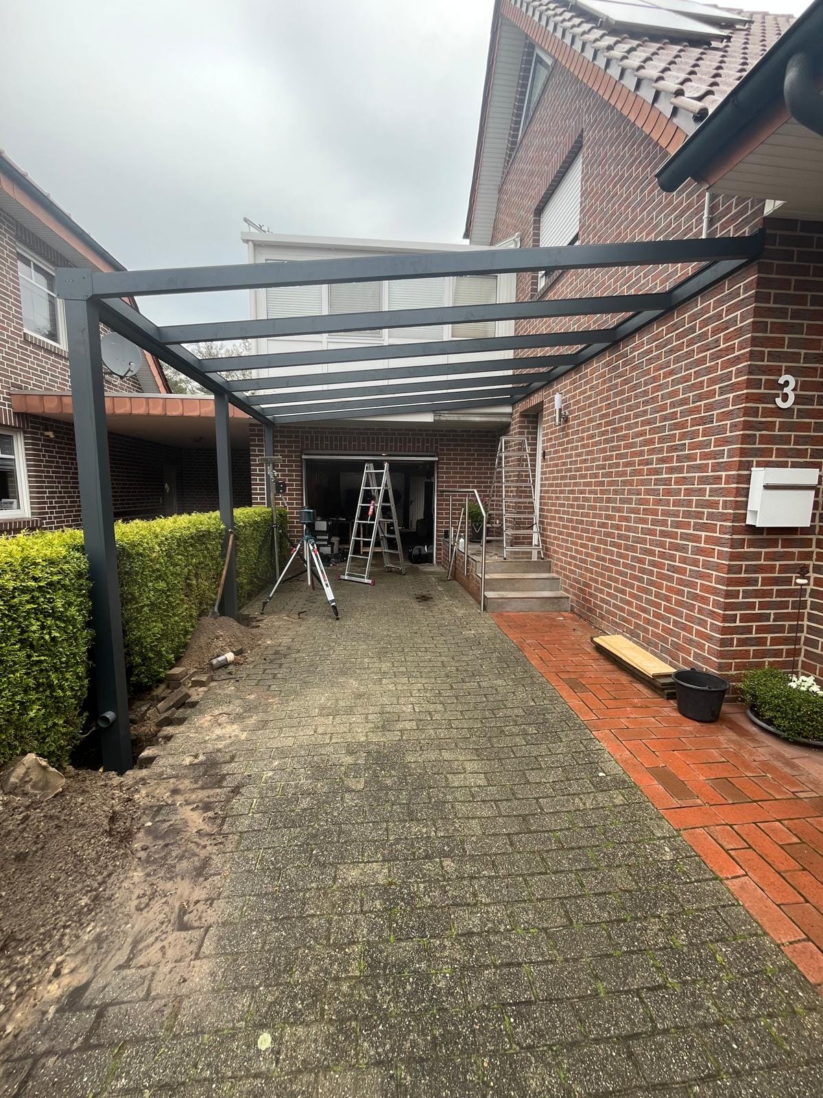 Anthrazitfarbener Aluminium-Carport mit Klarglasdach über der Einfahrt eines Klinkerhauses in Meppen-Schwefingen, aufgenommen während der Montage