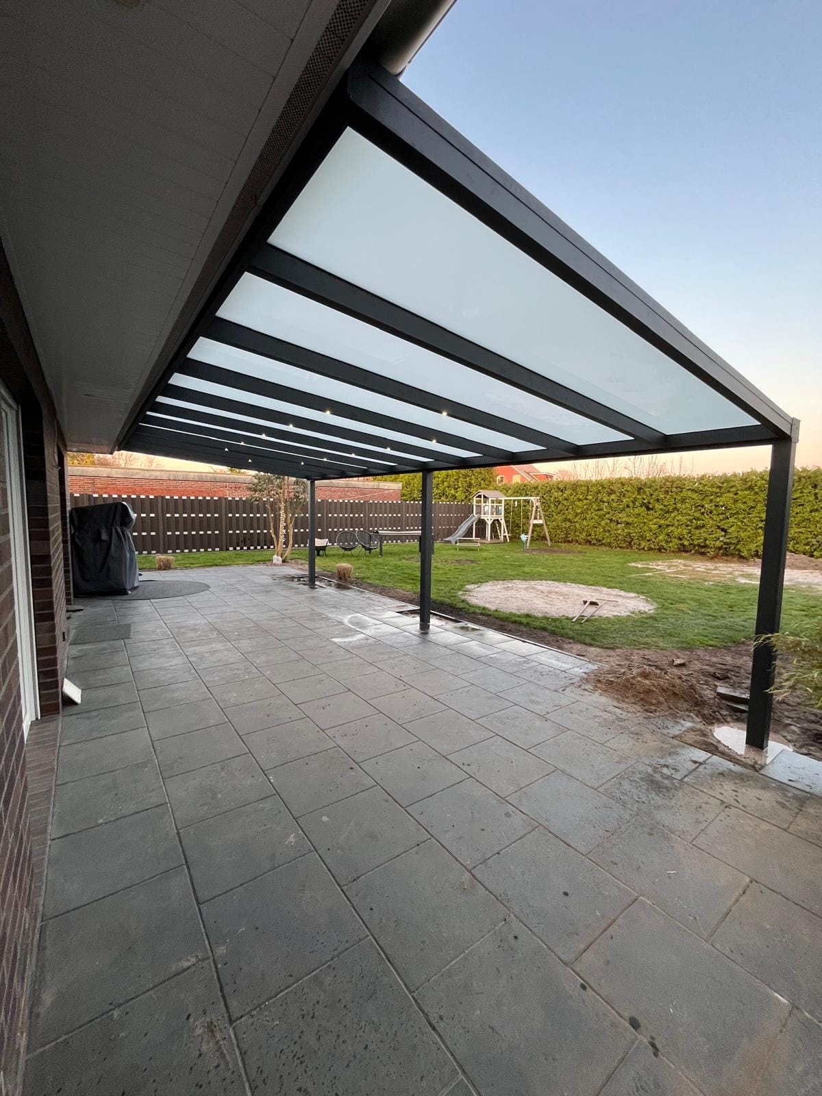 Basic-Überdachung in Anthrazit mit heller Dacheindeckung und LED-Spots in Apen, Blick über die Terrasse in den Garten mit Spielgeräten im Abendlicht