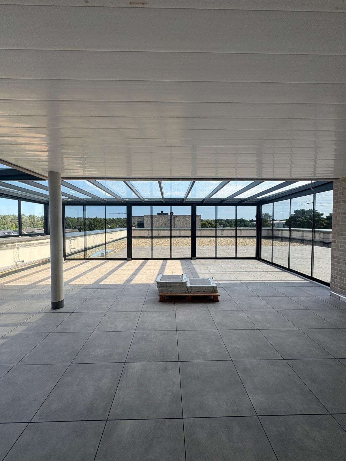 Blick von innen durch die getönten Glasschiebewände der Cube-Überdachung in Lingen auf die Dachterrasse, darüber das klare Glasdach mit Sparren