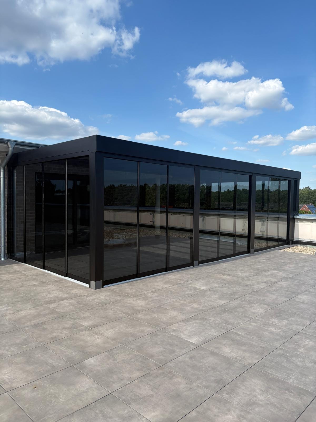 Schwarze Cube-Überdachung mit umlaufender Blende und getönten Glasschiebewänden auf einer Dachterrasse in Lingen, dahinter heller Klinker und Kiesdach