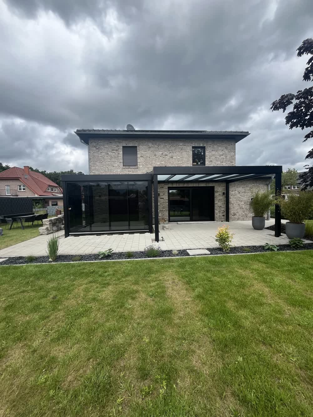 Schwarze Konstruktion an einem hellen Klinker-Neubau: links geschlossener Kaltwintergarten, rechts offene Terrassenüberdachung