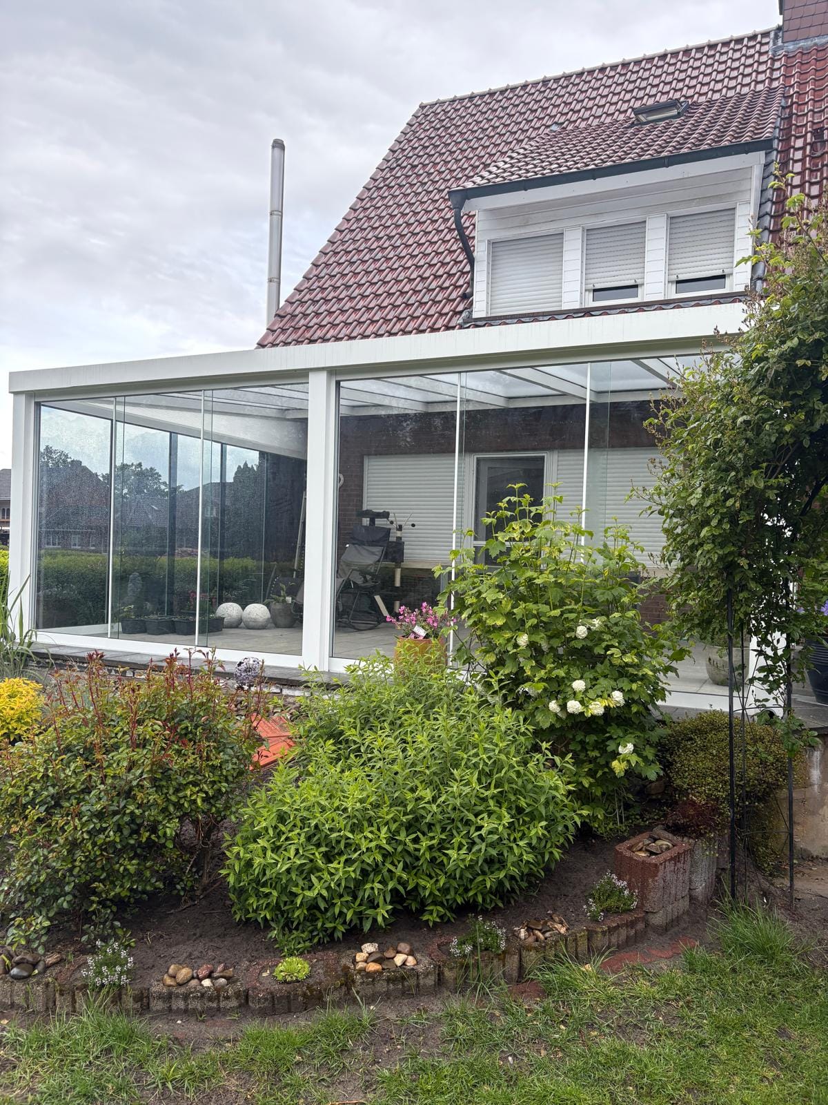 Weißer Kaltwintergarten mit Glasschiebewänden an einem Wohnhaus mit rotem Ziegeldach in Rütenbrock bei Haren, davor ein bepflanztes Beet