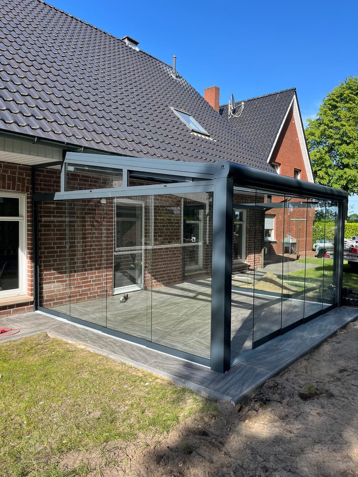 Anthrazitfarbener Kaltwintergarten in Wesuwe mit rahmenlosen Glasschiebewänden über Eck und keilförmigen Oberlichtern unter der Dachschräge