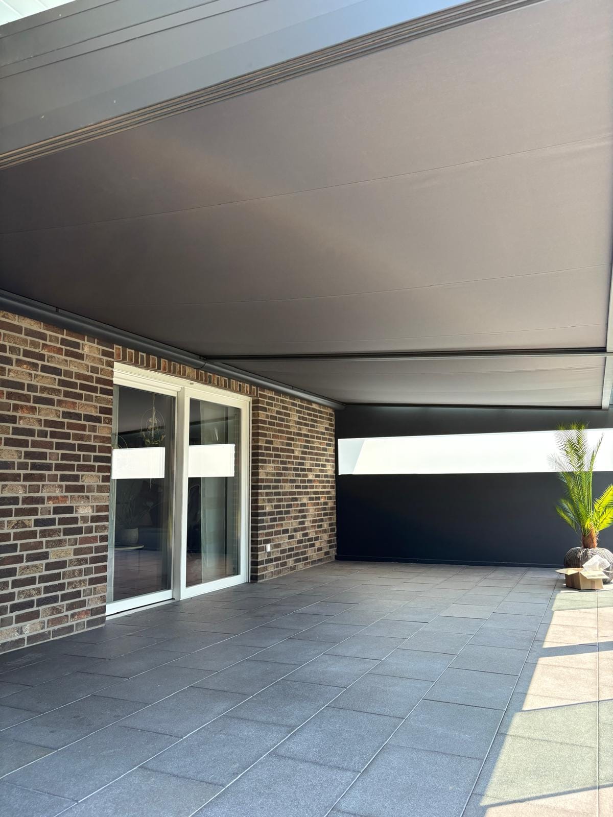 Ausgefahrene Unterglasmarkise in Taupe unter einer Terrassenüberdachung in Adorf bei Twist, darunter dunkelgraue großformatige Platten und Klinkerwand
